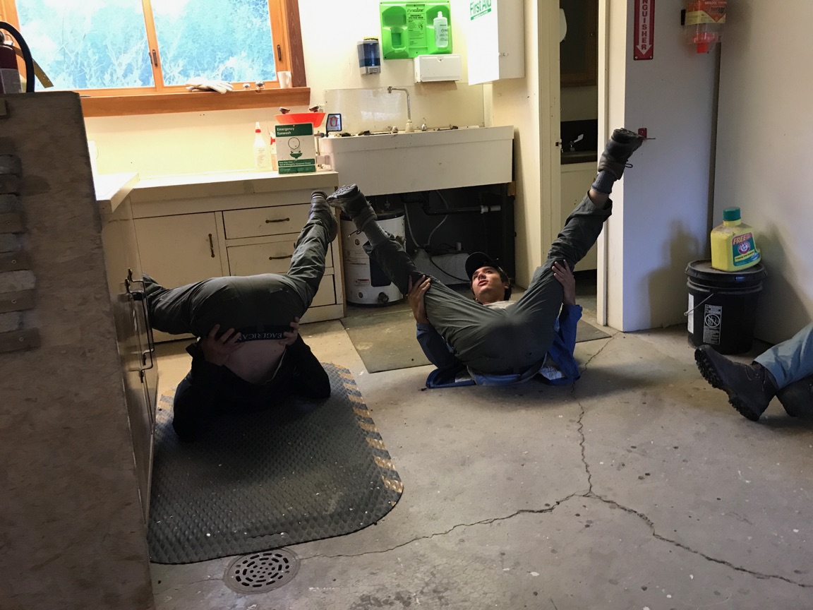 Two mechanics lying on their backs under a car in a workshop, laughing and lifting their legs up in the air.