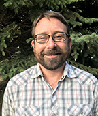 A bearded man in a plaid shirt standing in front of a tree.