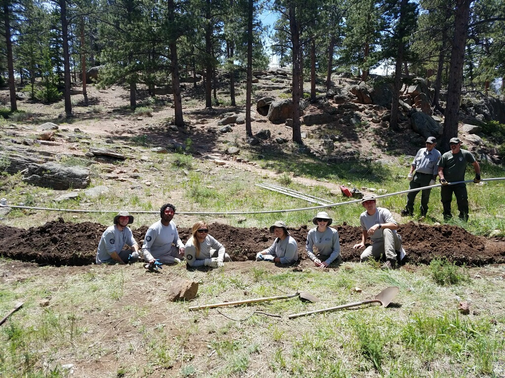 Group of eight people in work attire posing with shovels near a dug up area in a forested setting.