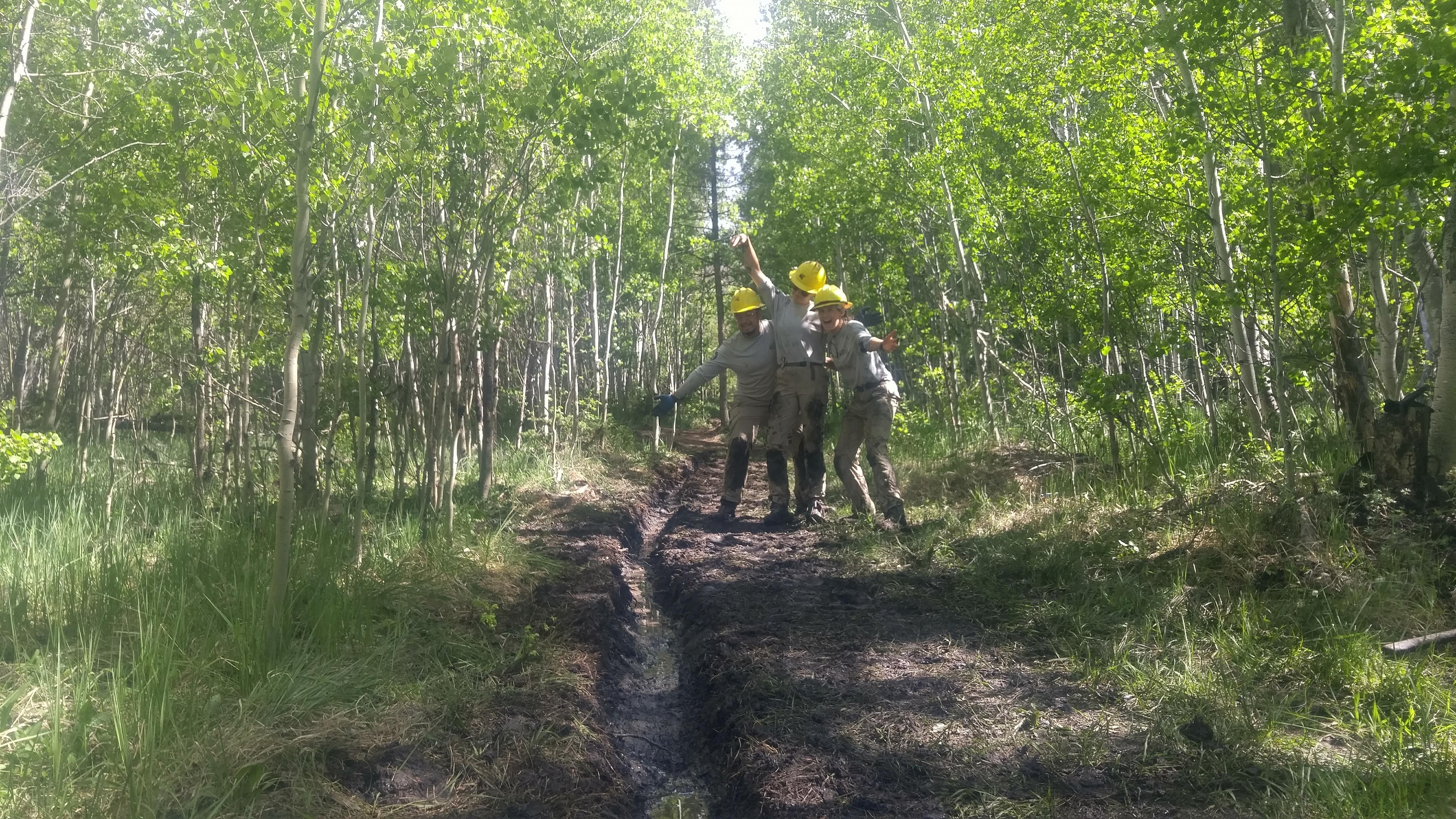 Two people wearing hard hats and work clothes surveying a muddy trail in a dense forest.