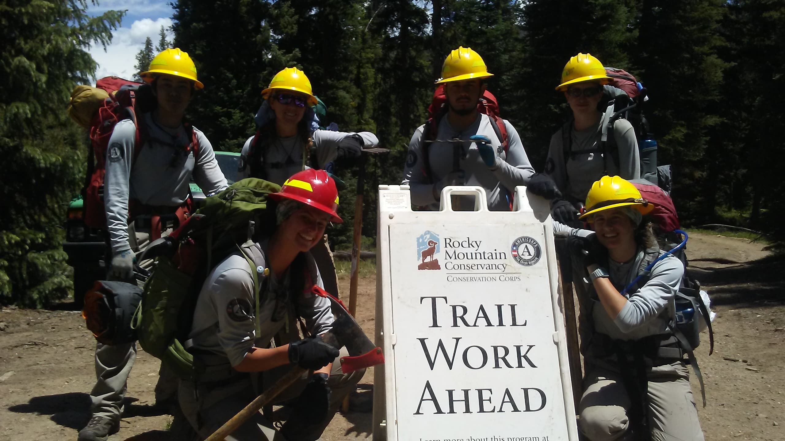 A group of seven conservation workers posing around a "trail work ahead" sign in a forest.