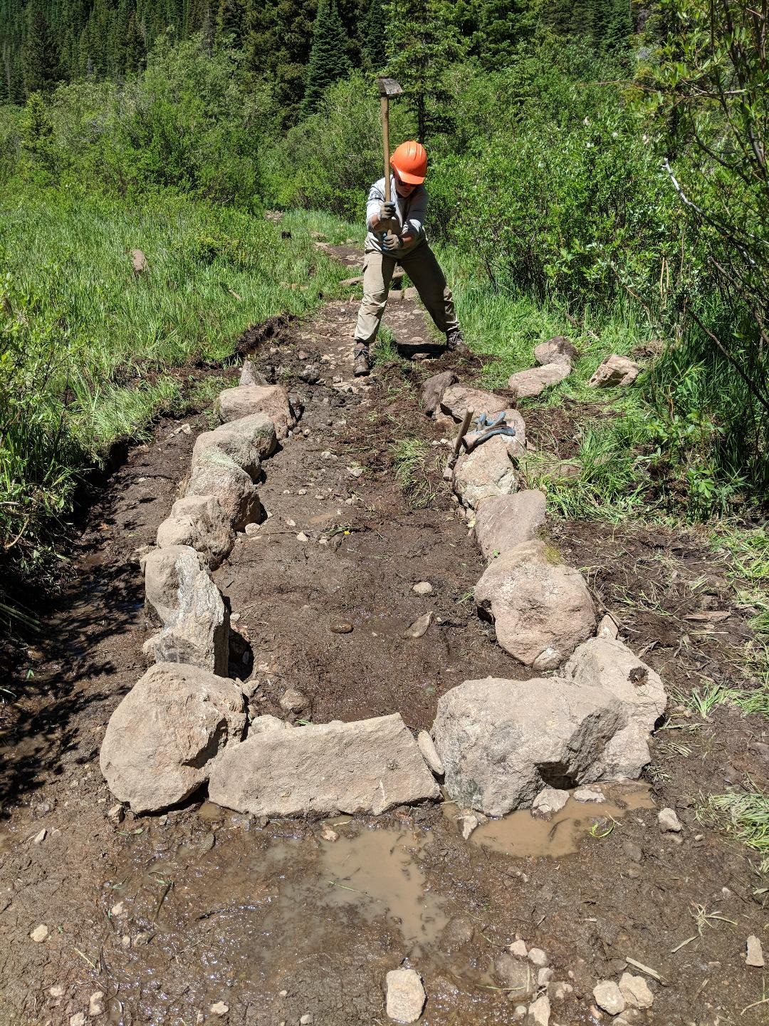 A person stands in a rocky, muddy trail surrounded by lush greenery.