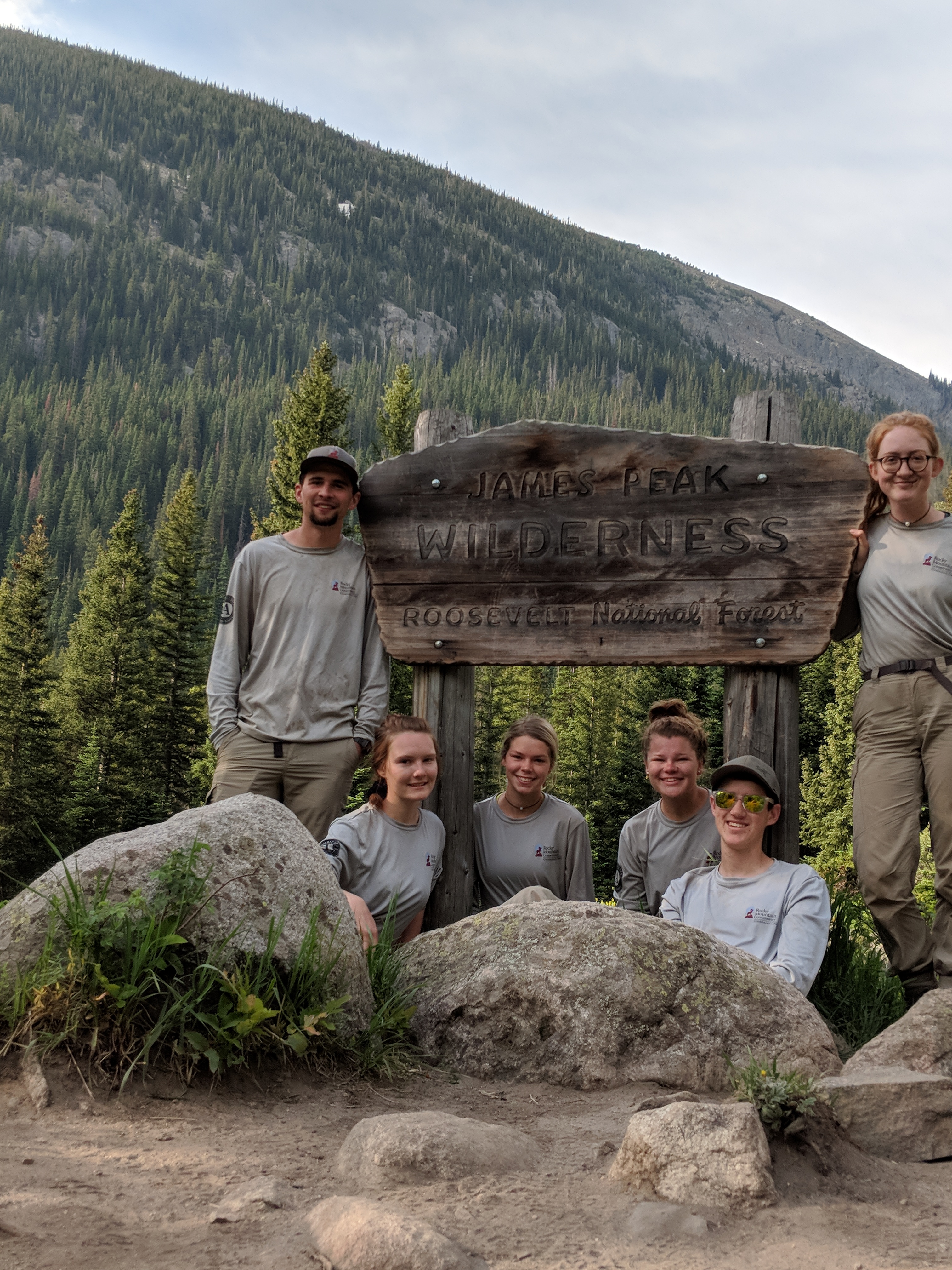 A hikers smiling in front of a wooden sign, surrounded by forested mountains.