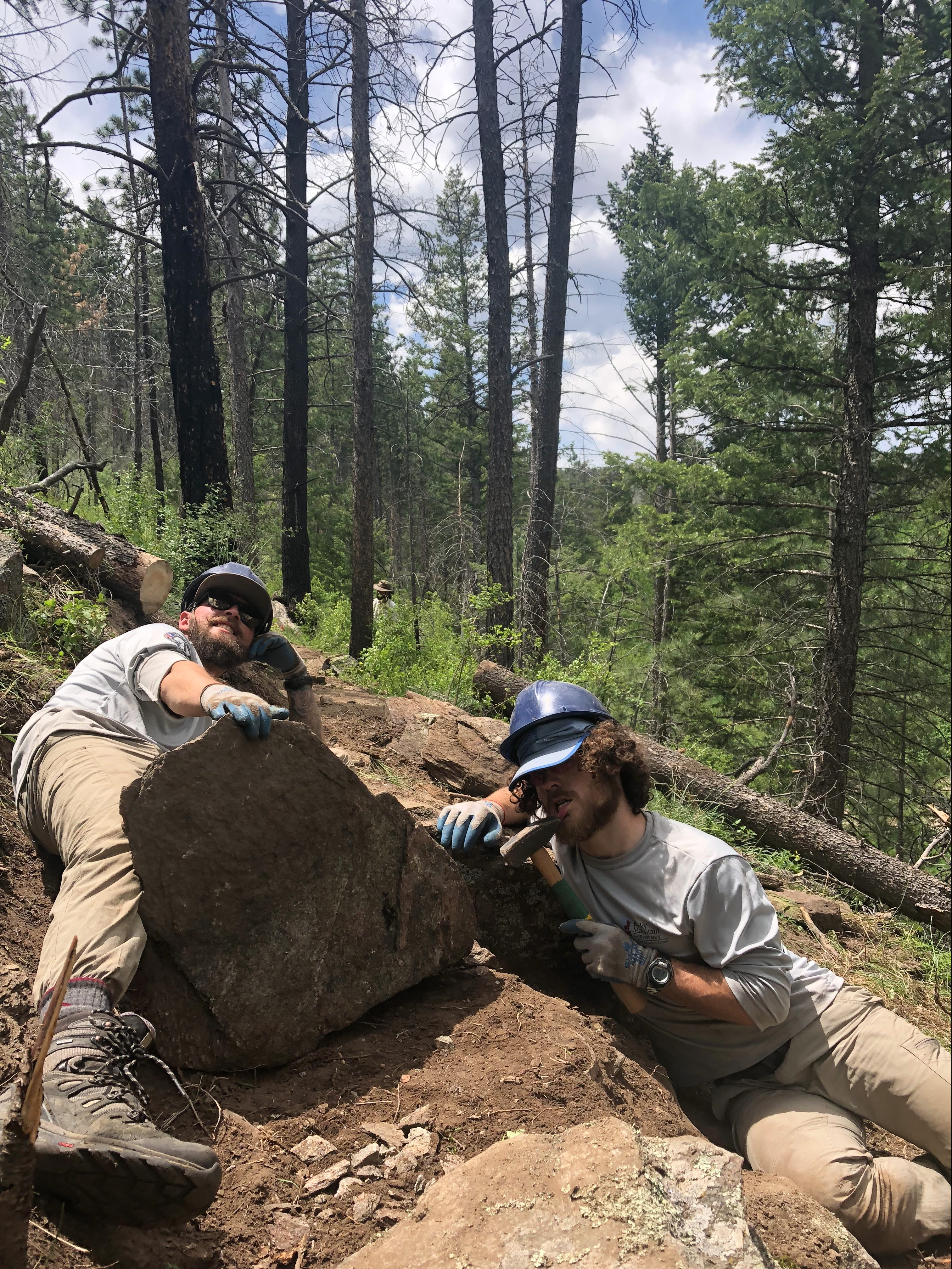 Two workers in helmets and gloves pose beside a large rock on a forested hillside