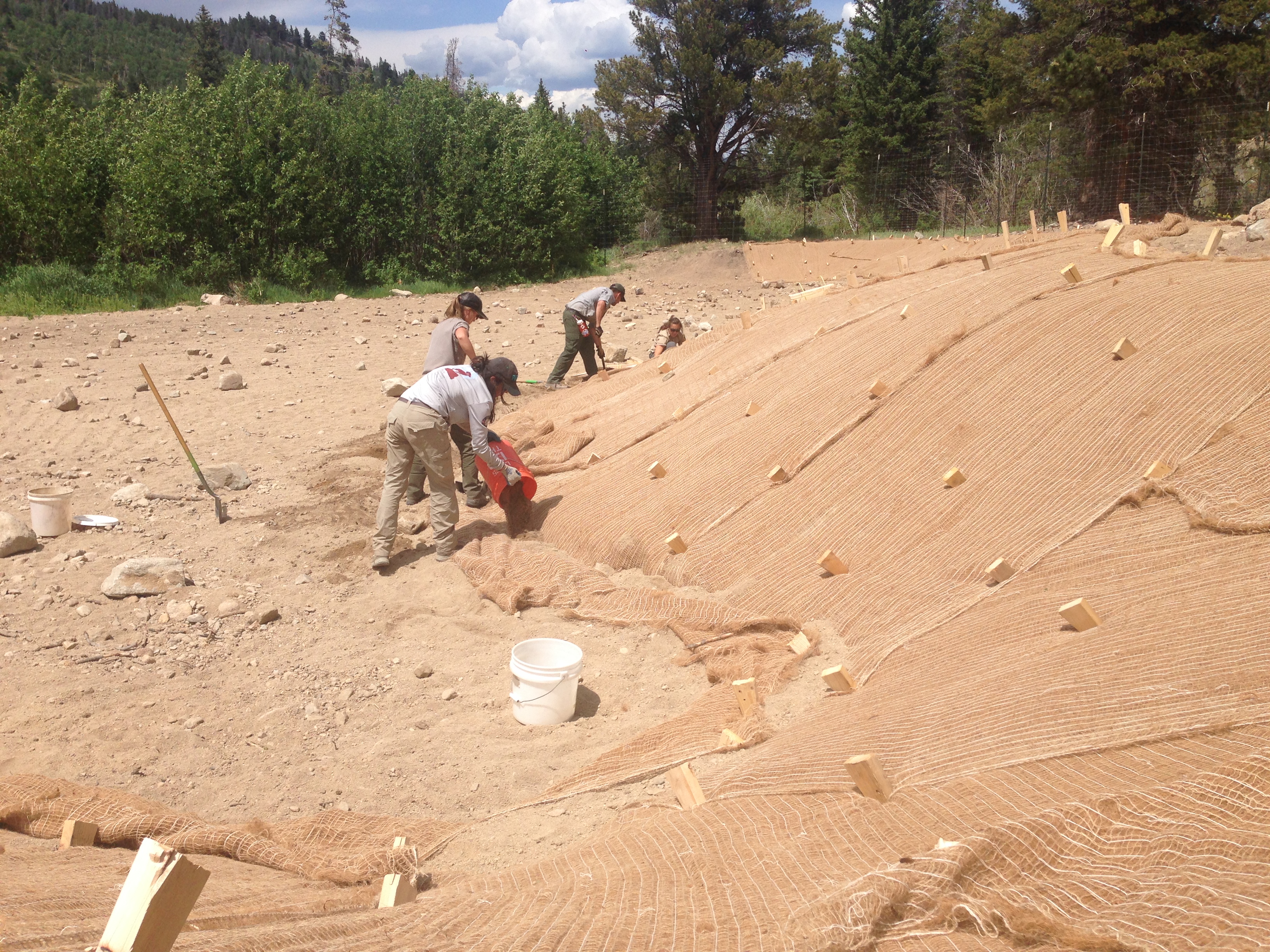 Workers install erosion control blankets on a sloped, sandy terrain