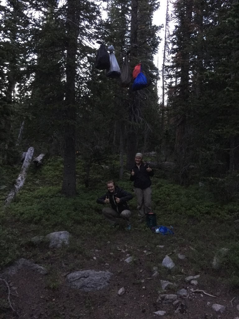 Two hikers giving thumbs up under their suspended backpacks in a forest at dusk.