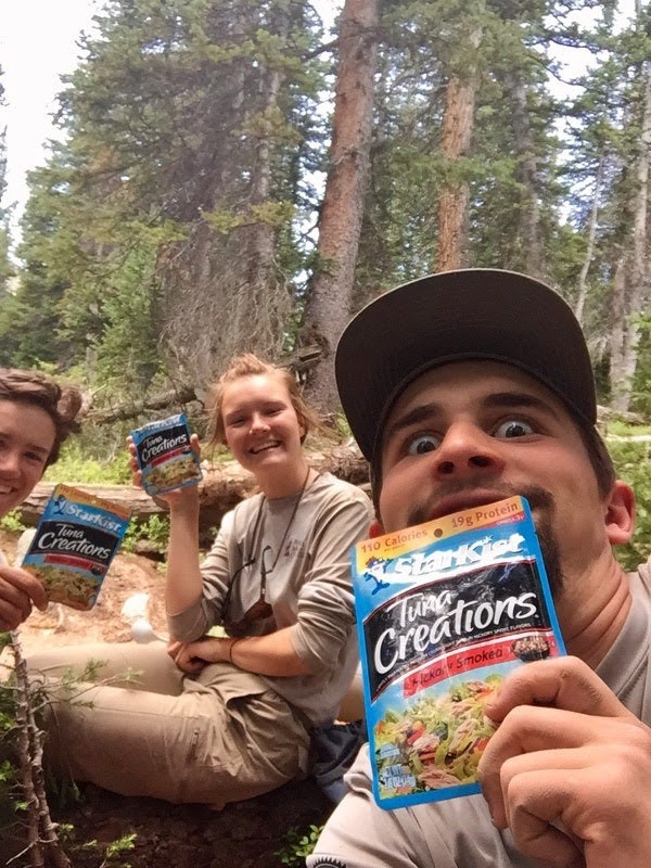 Three hikers hold up canned tuna products with excited expressions while sitting in a forest.