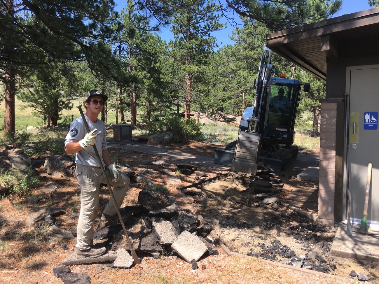 A park ranger stands holding a shovel near a digger clearing debris next to a park restroom surrounded by trees.