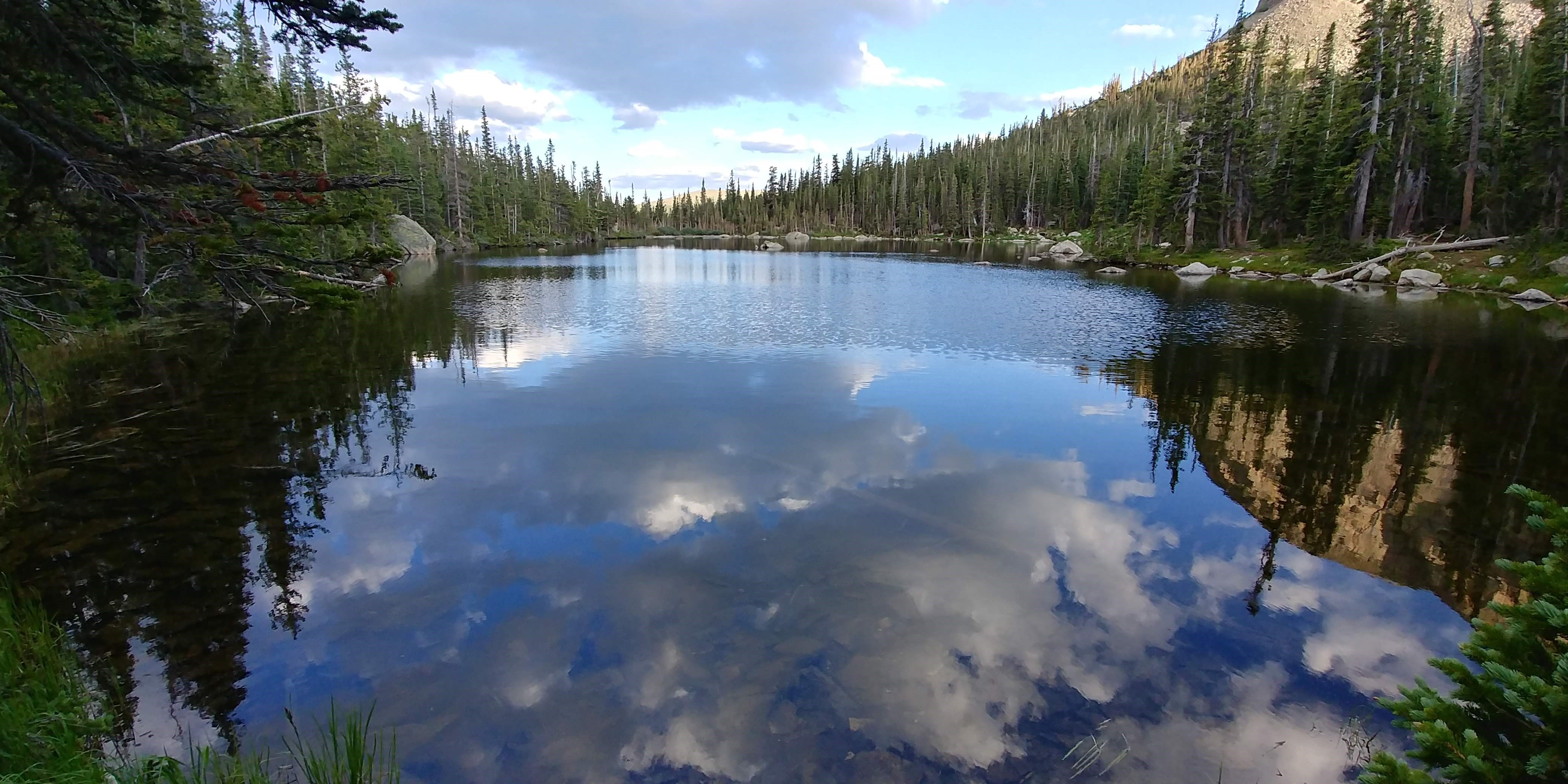 A serene mountain lake with clouds reflected in its still water, surrounded by dense coniferous forest under a partly cloudy sky.