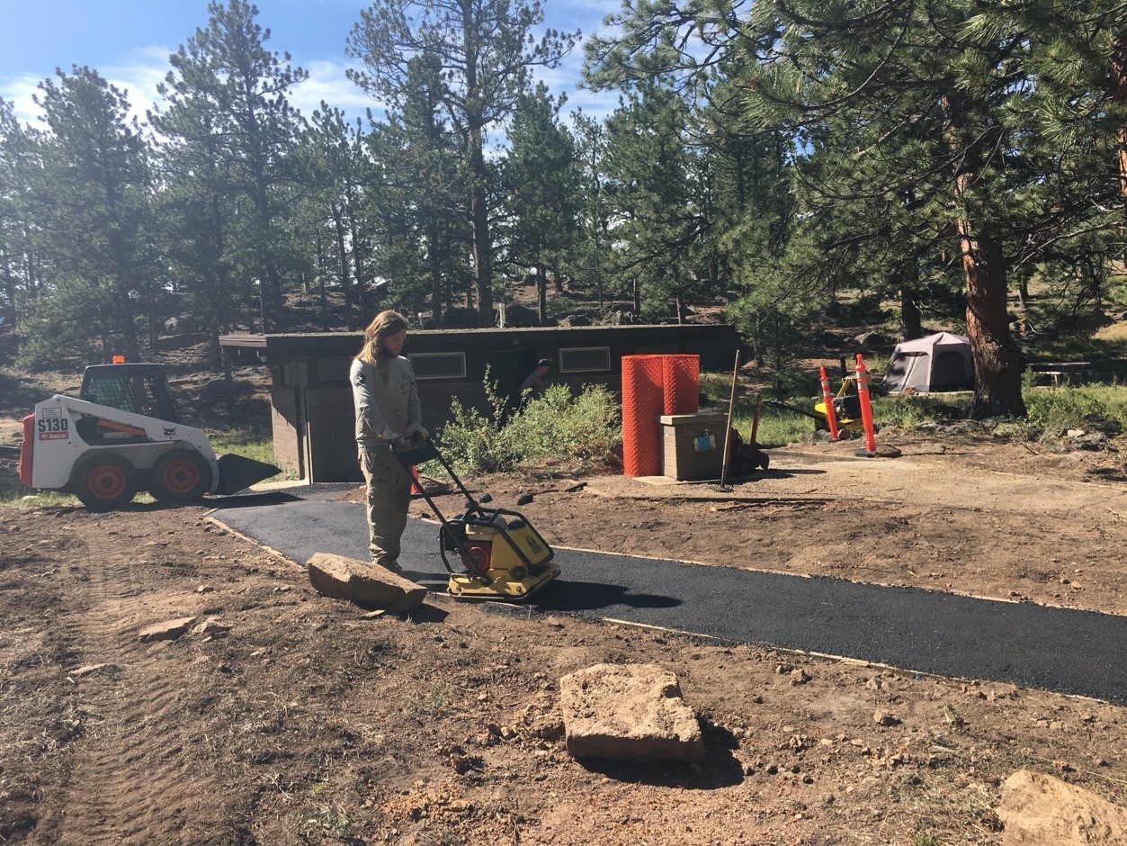 A person using a compacting machine on fresh asphalt in a forested area