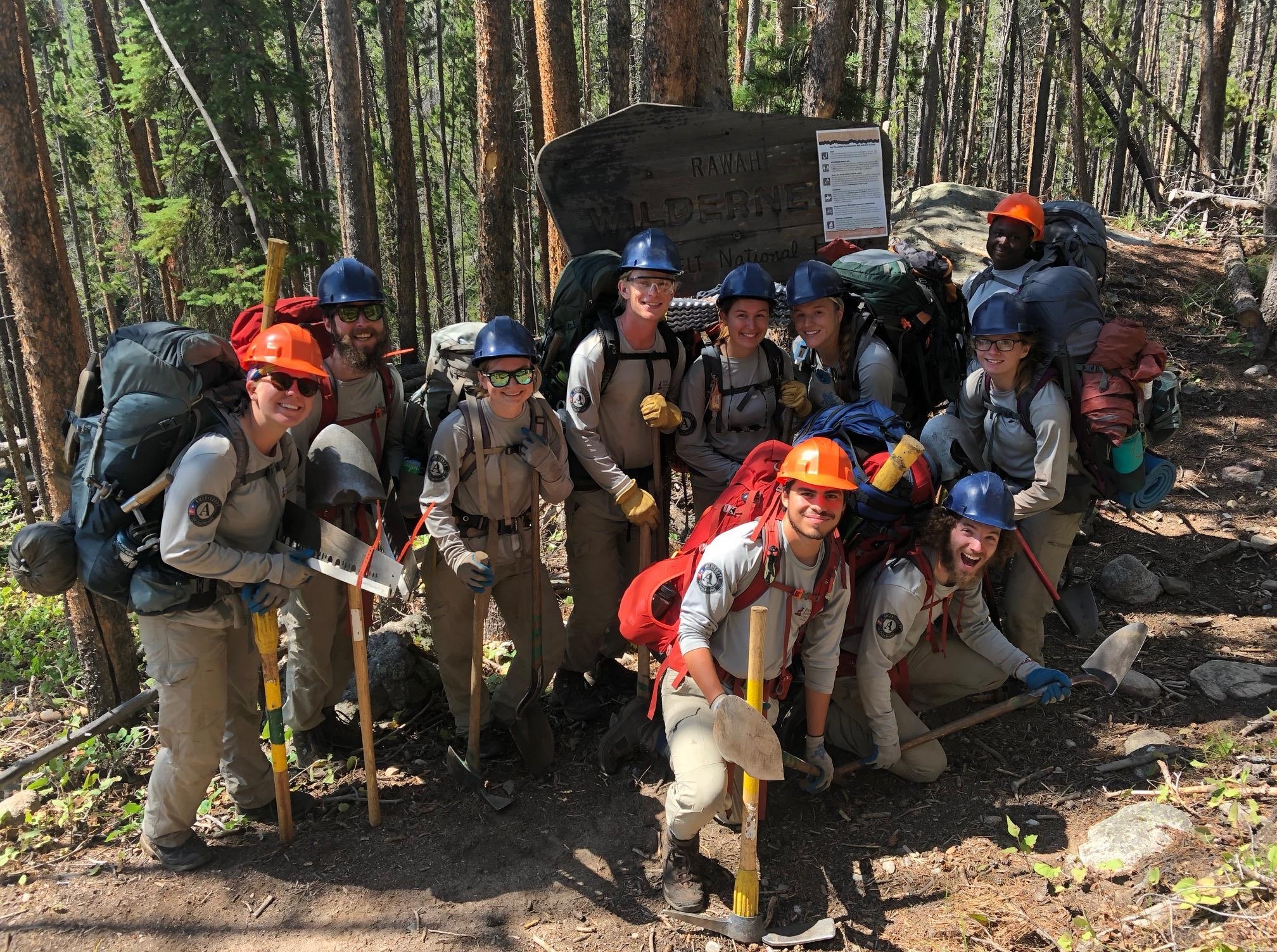 A group of forest rangers posing beside a wilderness sign in a forest.