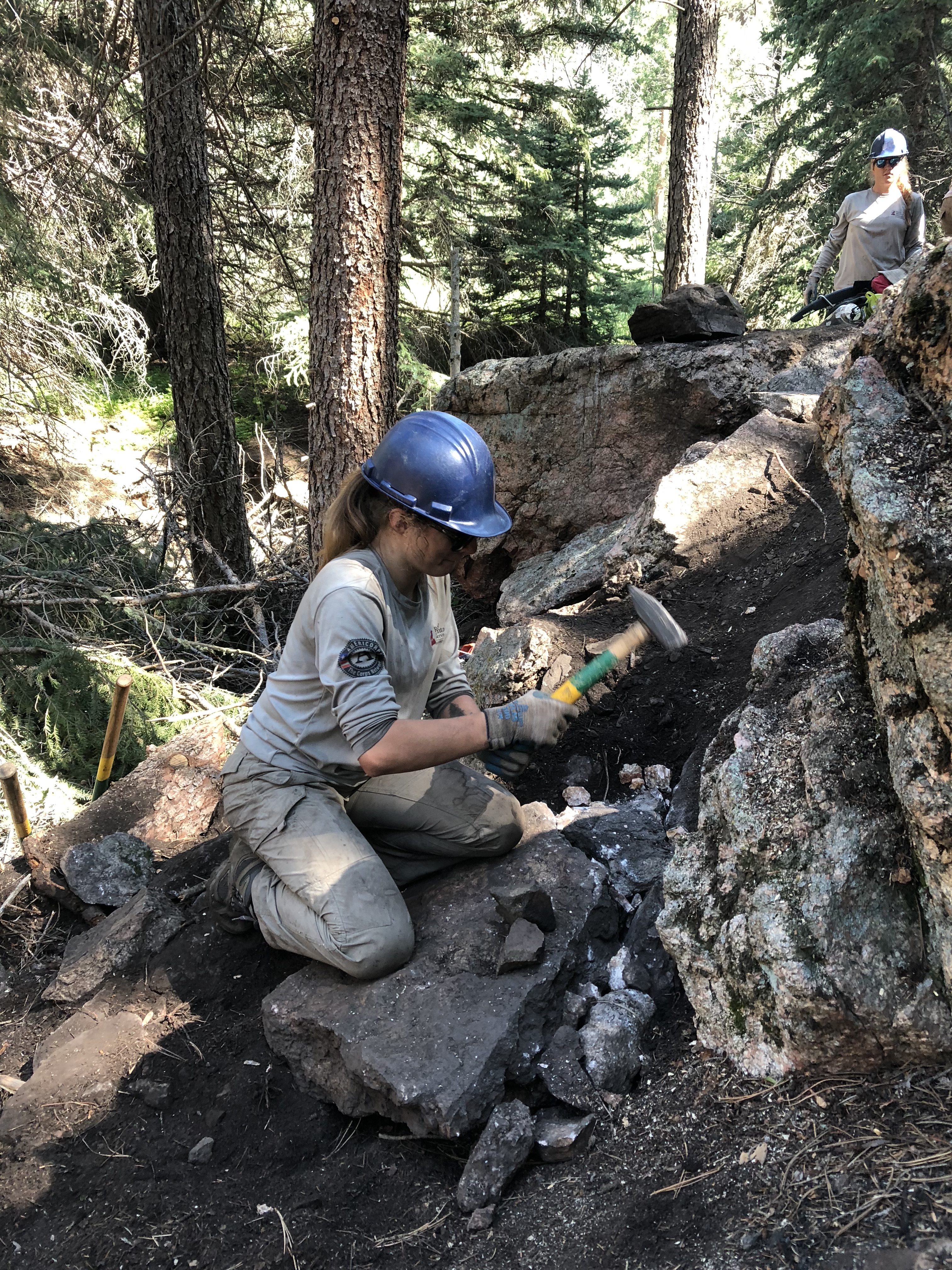 A worker in a hard hat uses a hammer to build a stone path in a forest, with another worker sitting nearby.