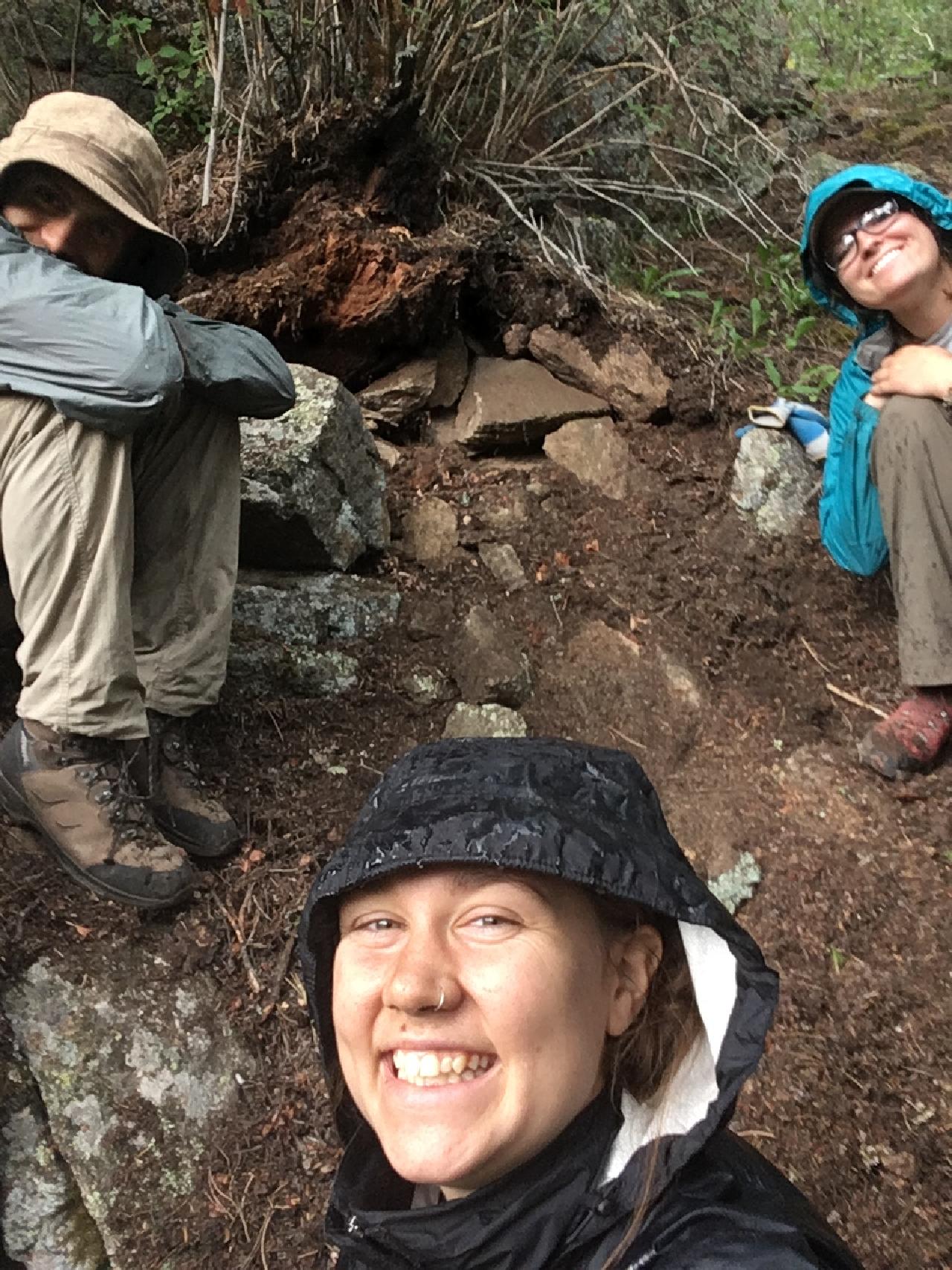 Three hikers taking a selfie, one in the foreground smiling with a rain hat; two others in the background, one covering face, sitting by a tree on a rocky trail.