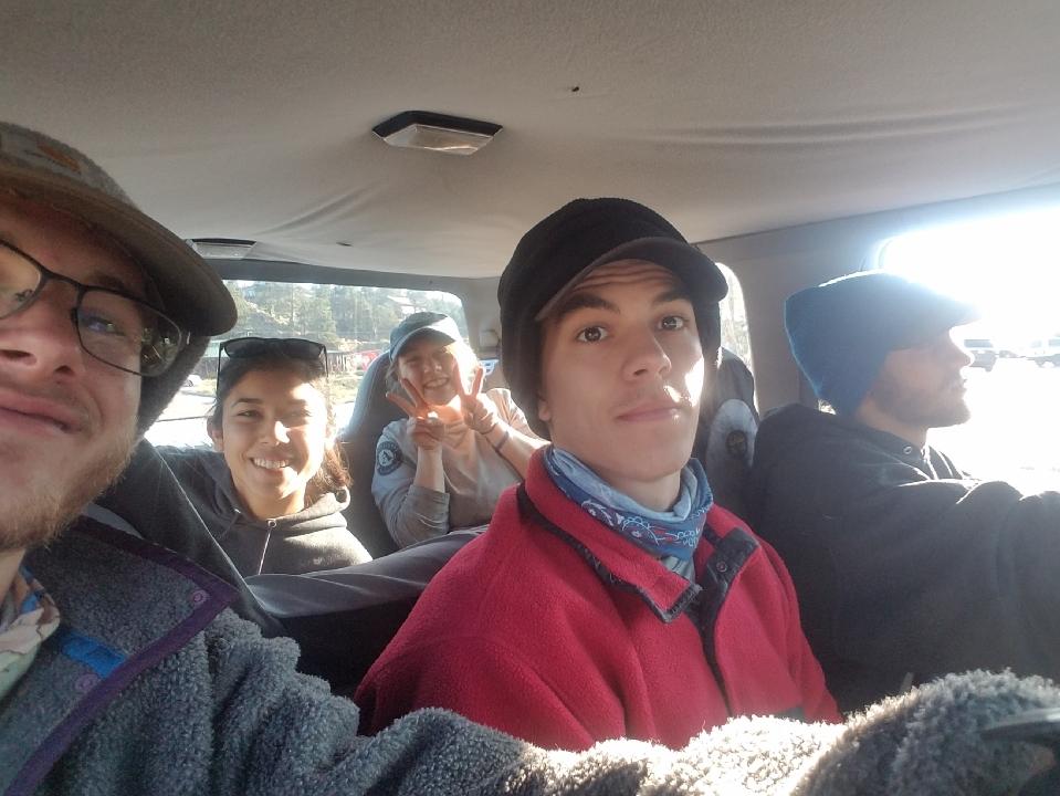 Five friends in a car, smiling at the camera; one person is driving, while the others pose from the passenger seats.