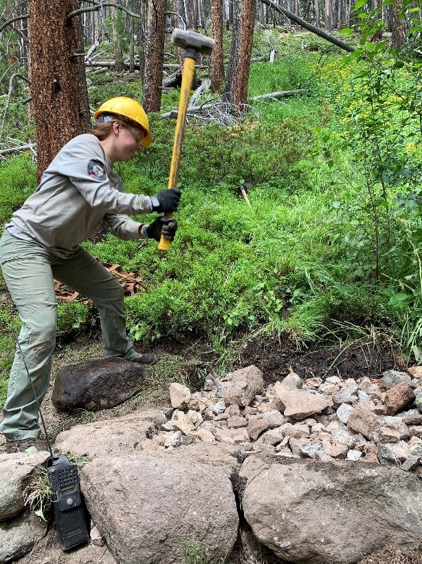 A woman is hammering rocks in a wooded area.