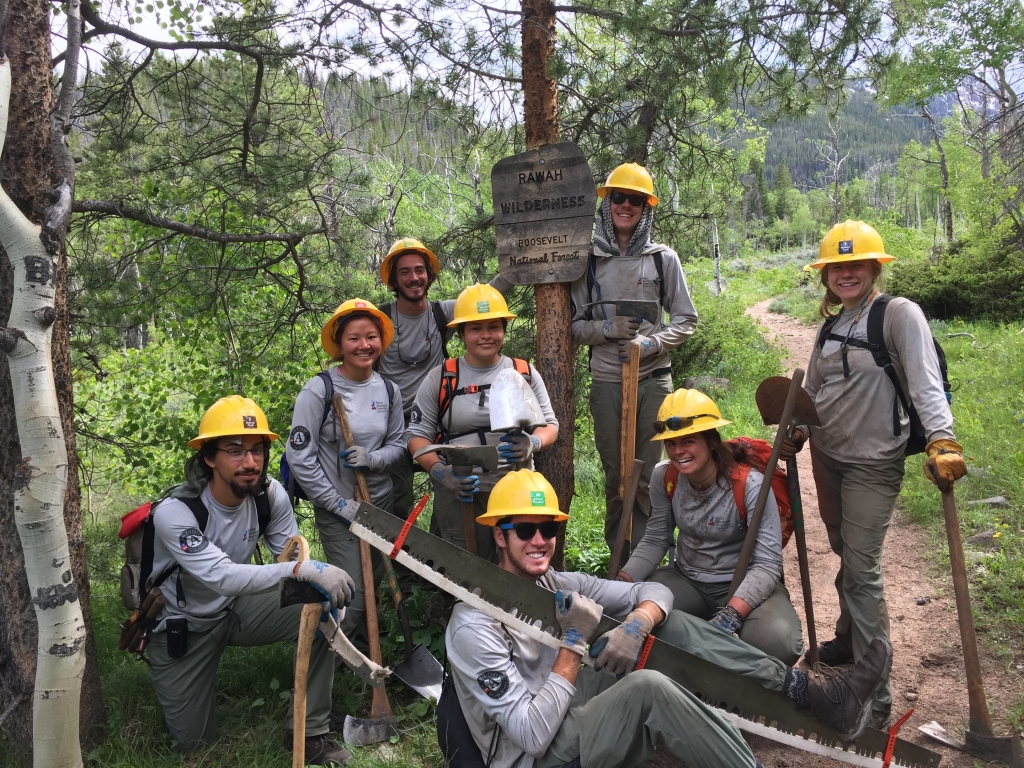 A group of forest workers posing with tools next to a wilderness trail sign