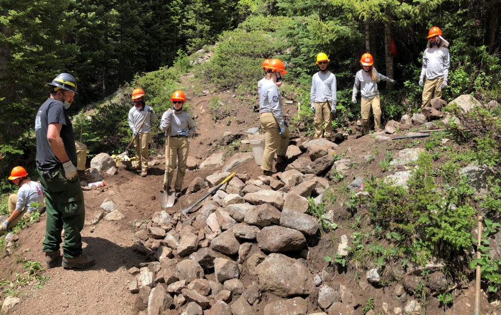 A group of workers in hard hats and protective gear constructing a rocky path in a forested area.