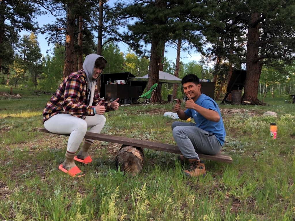 Two people sitting on a sawhorse in the woods.
