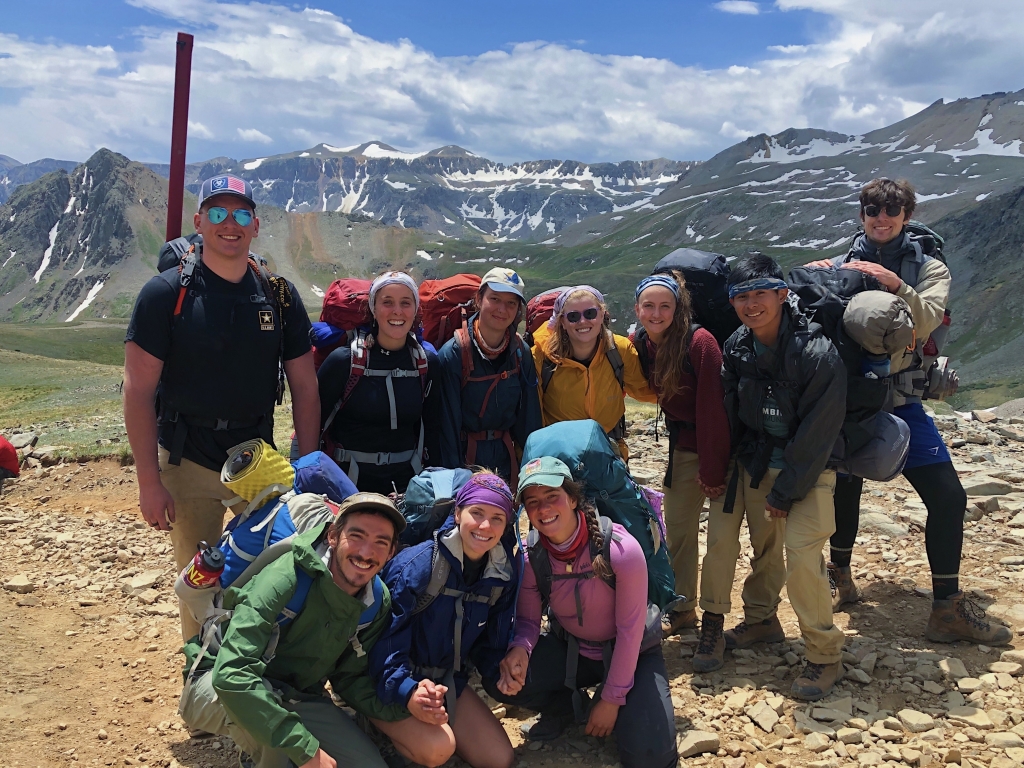 Group of nine hikers smiling at the camera on a mountain trail with snowy peaks in the background.