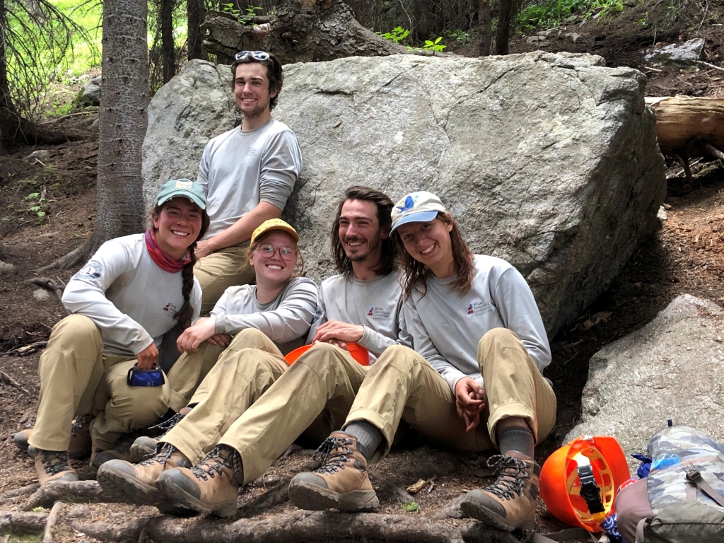 Group of smiling trail workers sitting on logs in a forest setting