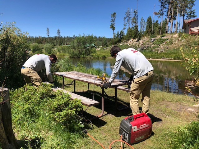 Two men working on a picnic table near a lake.