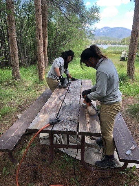 Two women working on a picnic table in the woods.