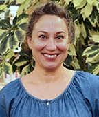 A smiling woman in a blue shirt standing in front of a plant.