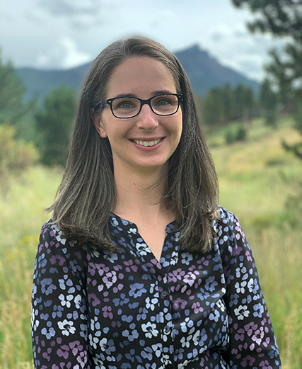 A woman in glasses standing in a field with mountains in the background.