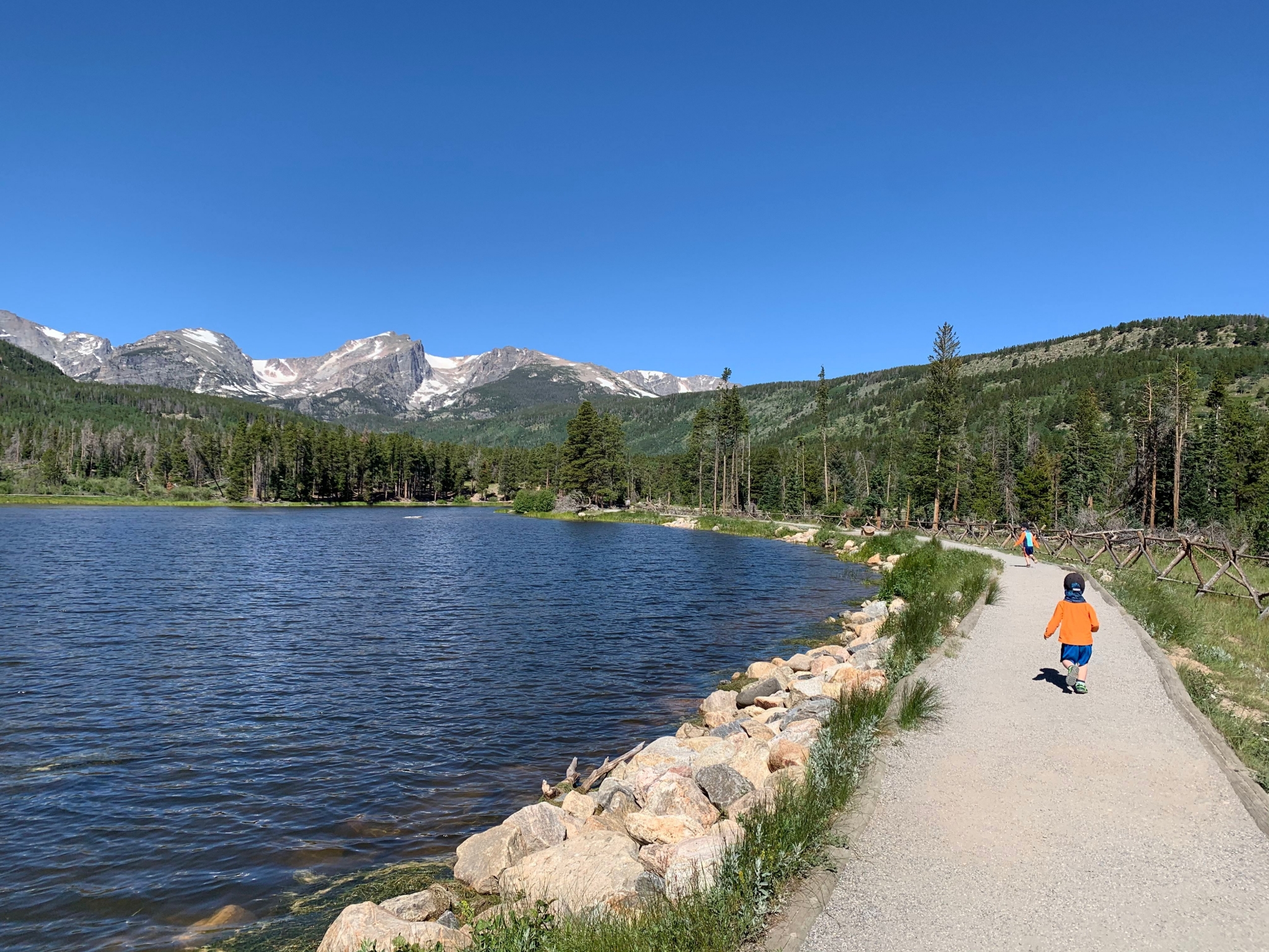 A child is walking along a path near a lake with mountains in the background.
