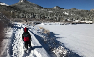 A child in black and red winter clothing and snowshoes walks away from the viewer on a snowy trail next to a snow-covered lake. Snowy, tree-covered mountains and blue skies are visible in the distance.