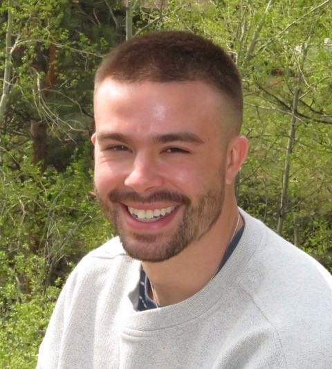 A man in a gray sweatshirt smiling in front of a tree.