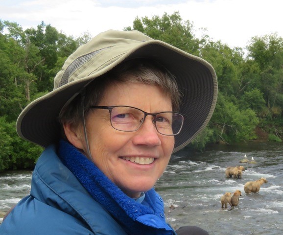 A woman wearing a hat and glasses in front of a river with bears.