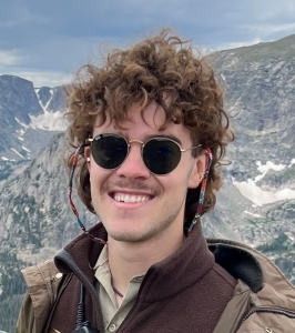 A smiling person with curly hair wearing round sunglasses and a jacket with a mountainous background.