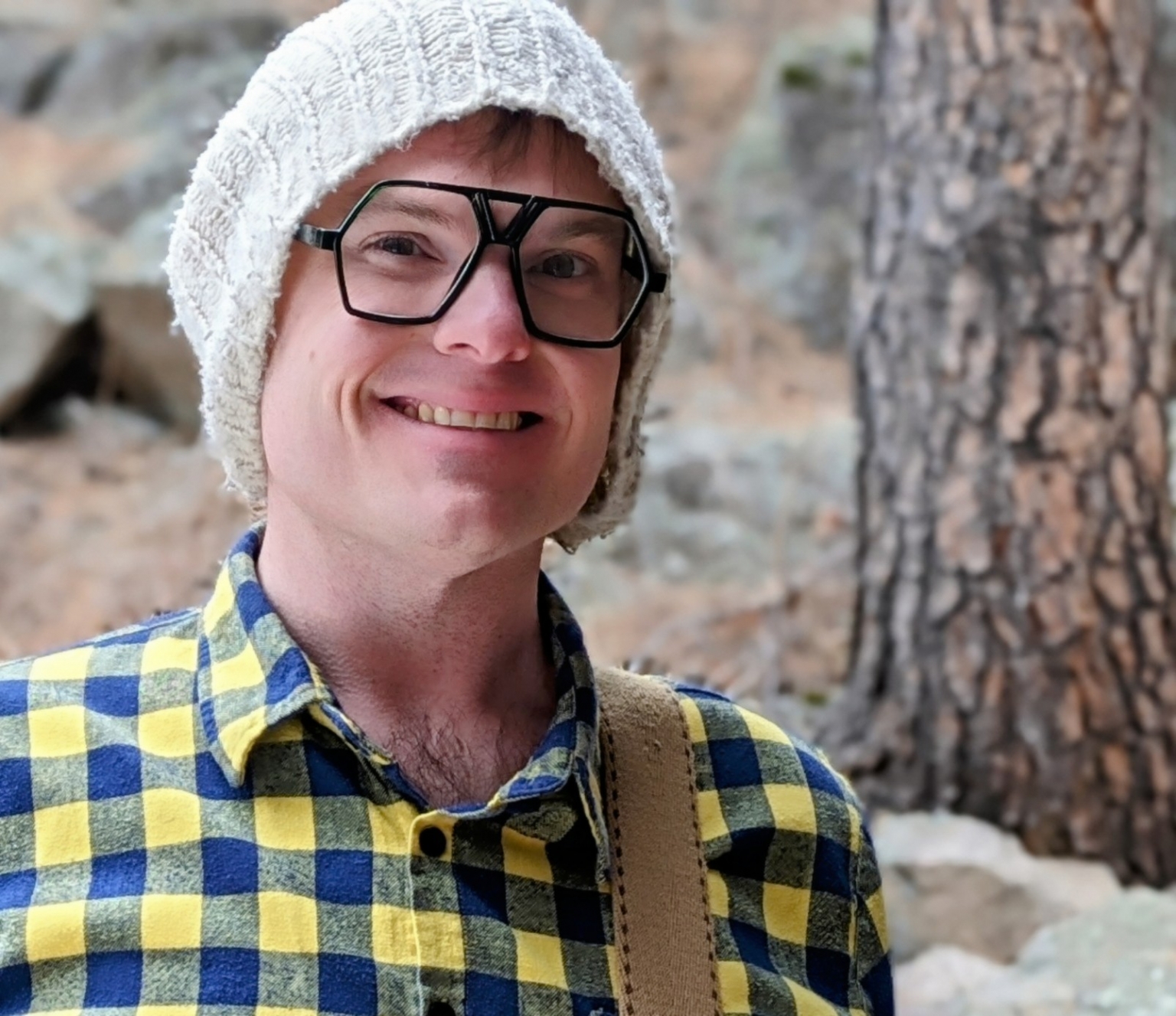 A man wearing glasses and a beanie is standing in the woods.