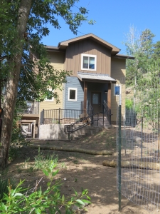 A two-story house with a brown and beige exterior, front steps, and a sloped yard surrounded by trees and fencing, set against a clear blue sky.