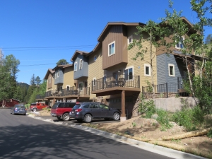 A row of modern, multi-story townhouses with garages and balconies, situated along a paved street with parked cars in front. Trees and a mountain are visible in the background.