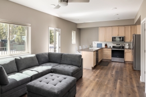 A modern, open-concept living room and kitchen with a gray sectional sofa, an island with barstools, wood cabinets, stainless steel appliances, and wood flooring. Light streams in through large windows.