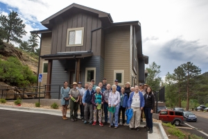 A group of people stands together in front of a two-story building on a paved area. Pine trees and parked cars are visible in the background.