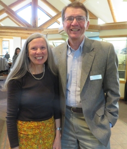 Two people are standing side by side indoors, both smiling at the camera. The woman on the left is wearing a dark top and patterned skirt. The man on the right is wearing a blazer and name tag.