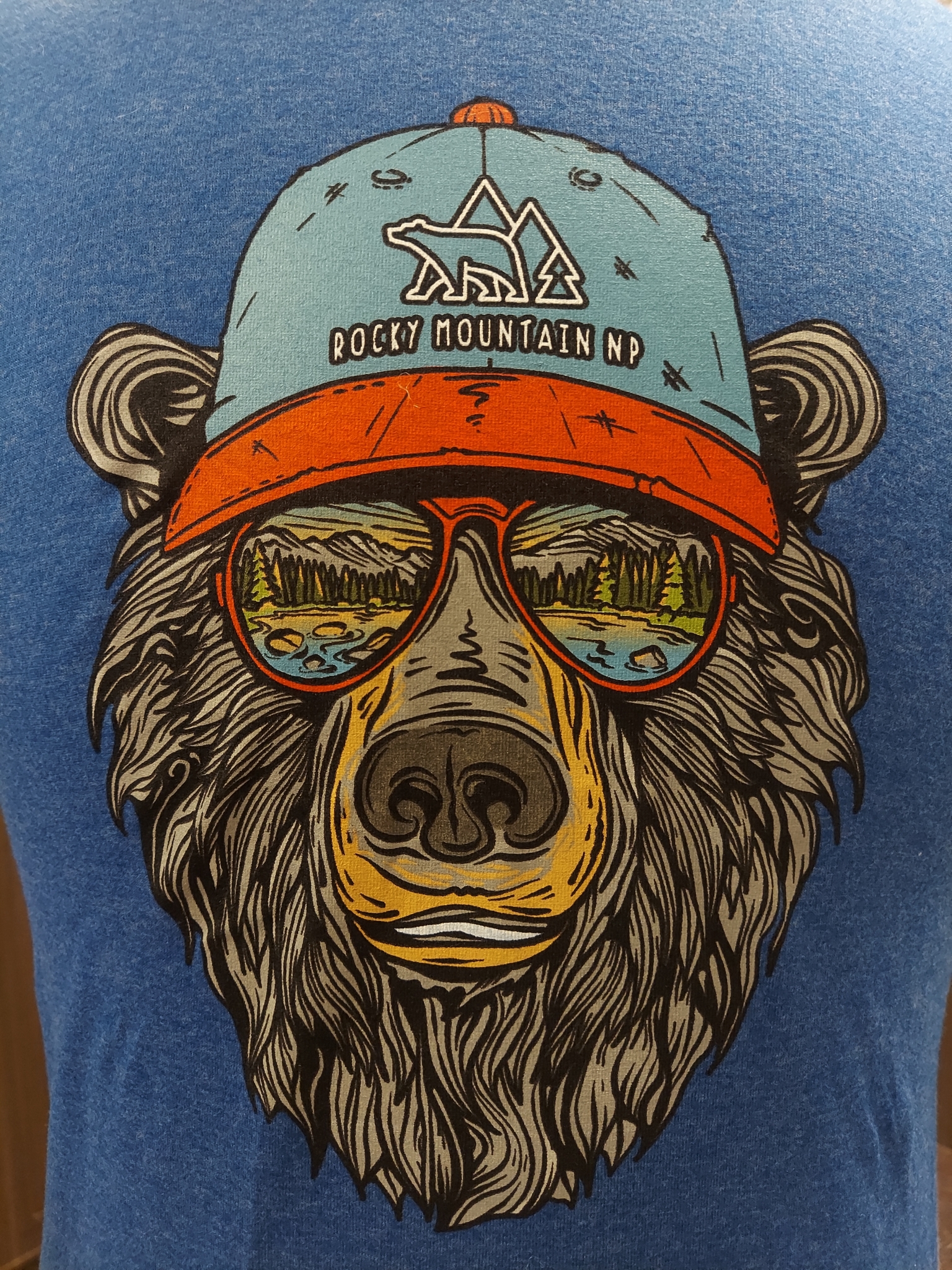 Product Description: This blue t-shirt features a graphic illustration of a bear wearing a cap that reads "Rocky Mountain NP" and sunglasses reflecting a scenic landscape, ideal for kids.