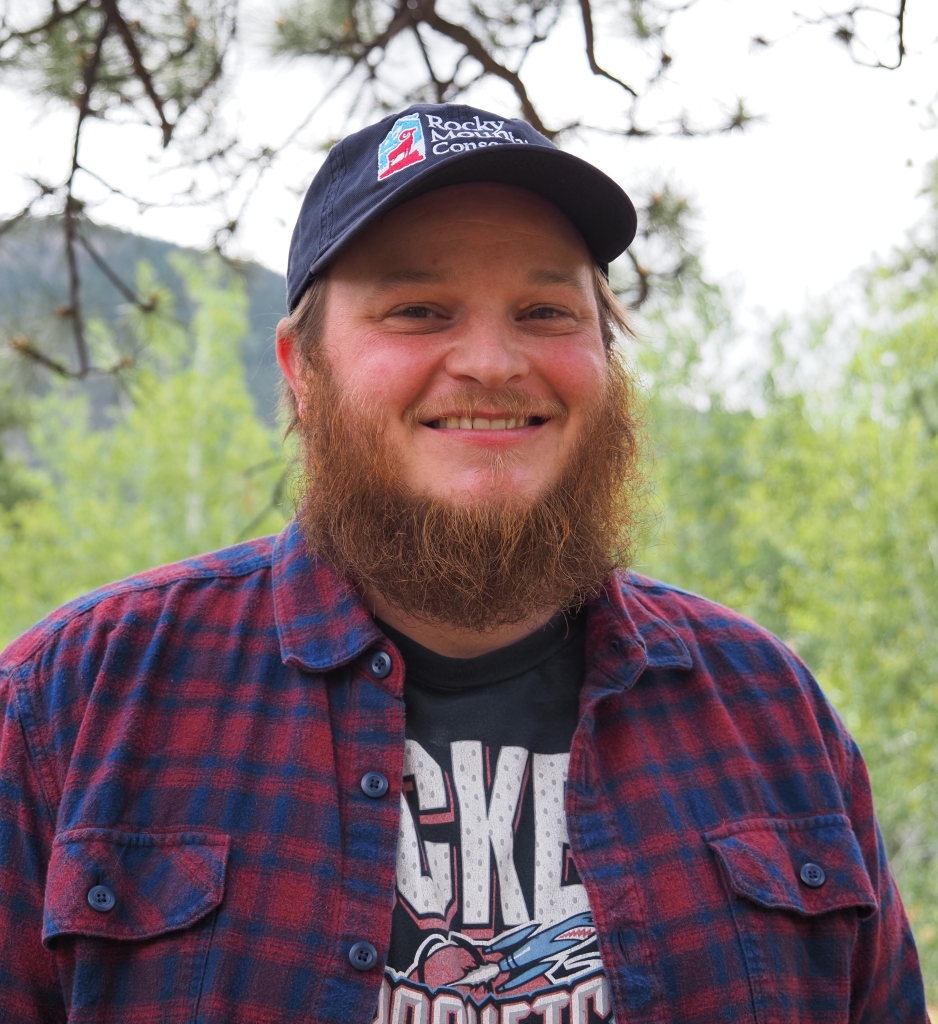 A person with a beard and a cap smiles at the camera. They are wearing a flannel shirt and a graphic T-shirt. Trees and a blurred natural background are visible behind them.