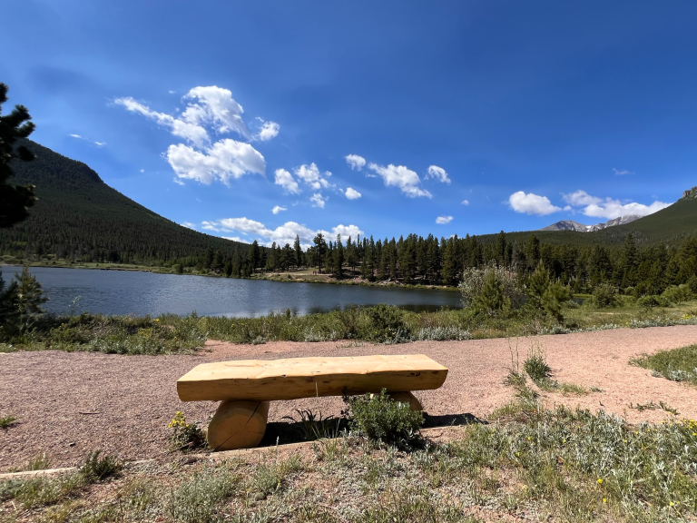 A wooden bench overlooks a serene lake surrounded by trees and hills under a blue sky with scattered clouds.