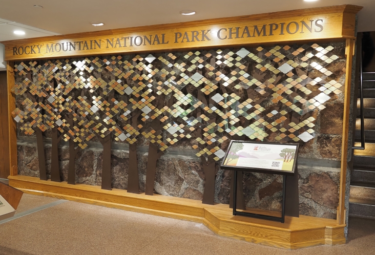 A display titled "Rocky Mountain National Park Champions" featuring a wall with tree designs and an array of small, square plaques, alongside an informational stand.