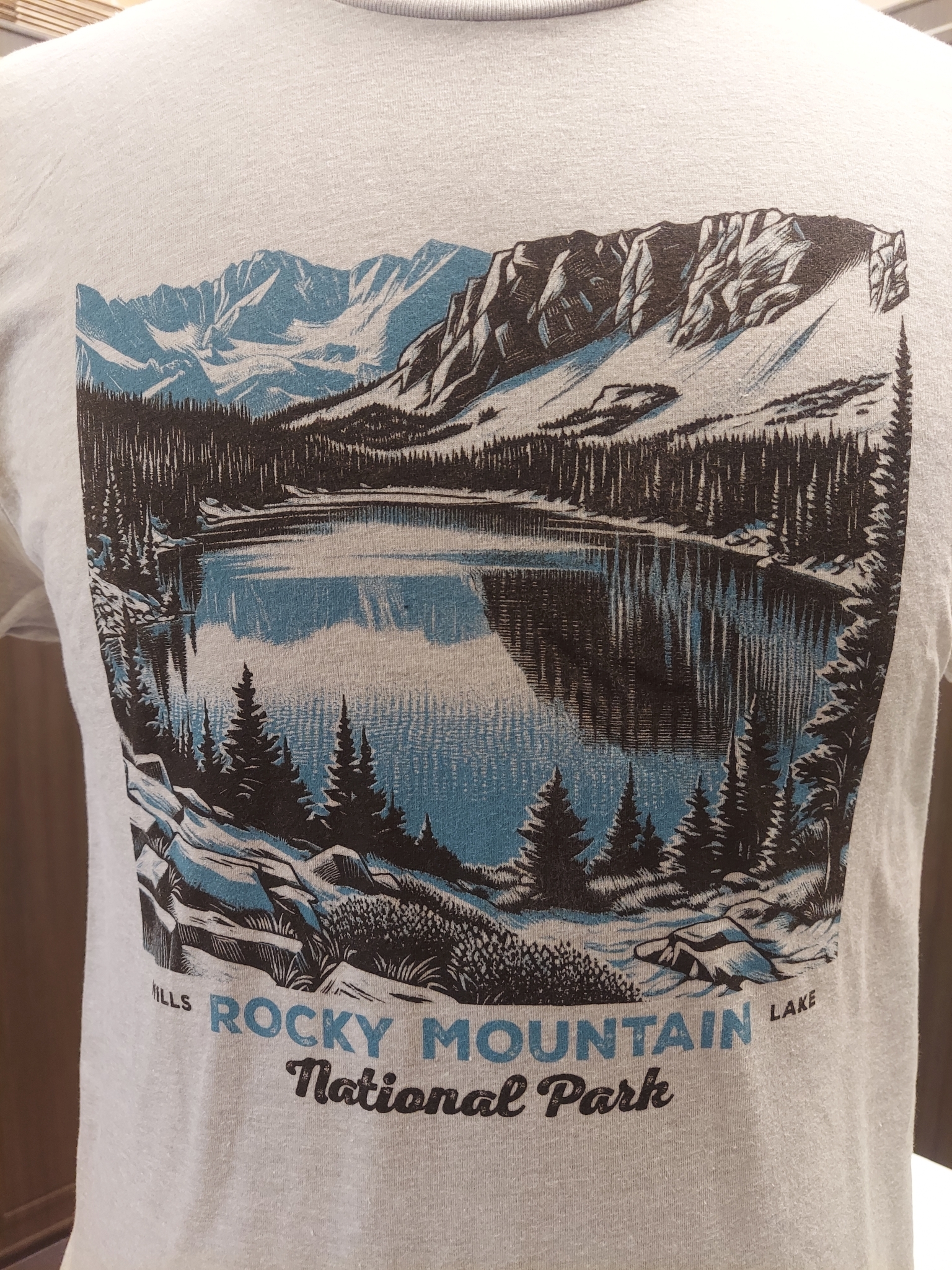 Close-up of the back of the T-Shirt - RMNP Mills Lake featuring an illustration of a lake surrounded by mountains and trees, with the text "Rocky Mountain National Park" at the bottom.