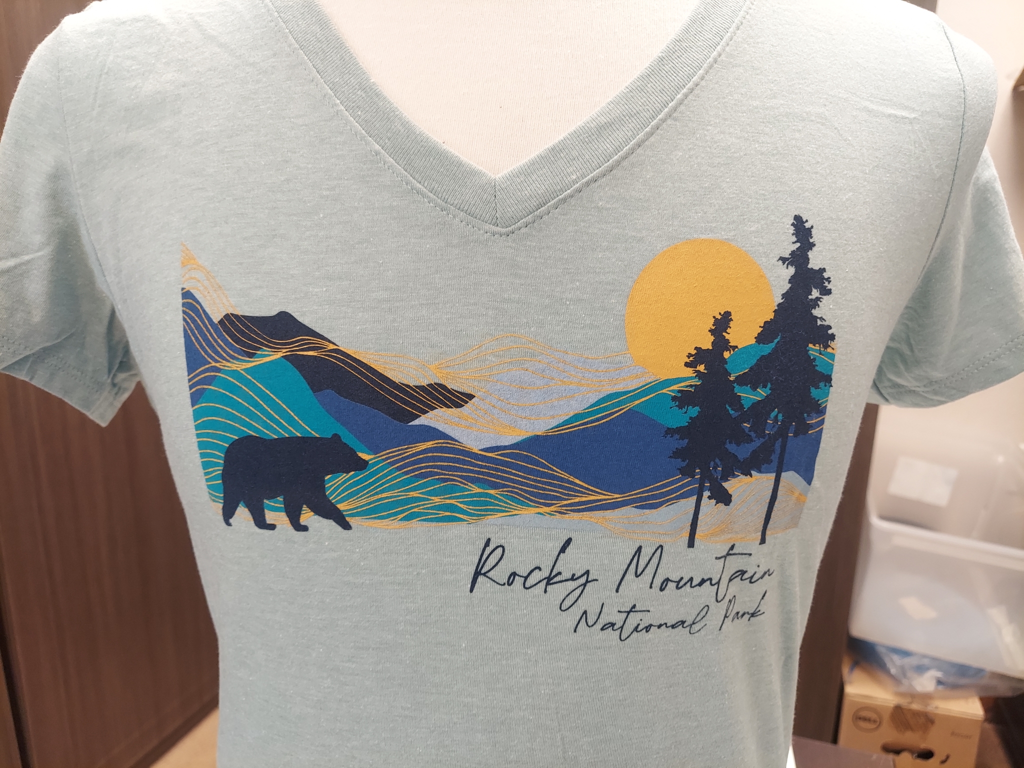 A T-Shirt - RMNP Bear Mist in light blue, featuring a bear, trees, mountains, and a sun design reminiscent of Rocky Mountain National Park.