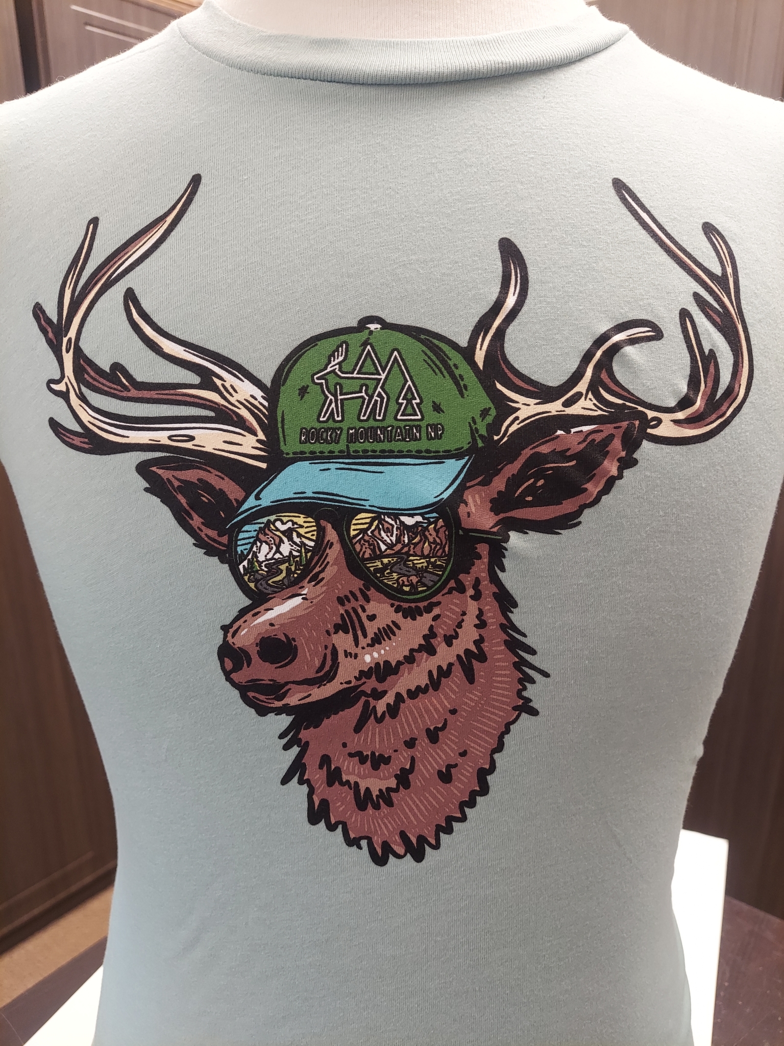 Illustration of a deer wearing a green cap and sunglasses, printed on the back of the light gray T-Shirt - Kids RMNP Elk Sunglasses.