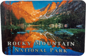 A photograph of Rocky Mountain National Park's Dream Lake, showcasing its tranquil waters bordered by rocky shores, lush forested areas, and majestic mountain peaks glowing in orange light. Text at the bottom reads, "Rocky Mountain National Park." Product Name: Magnet - RMNP Dream Lake