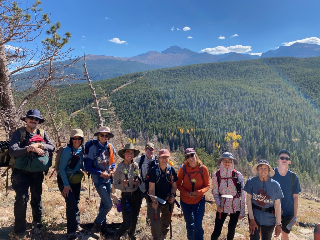 A group of people in hiking attire stand together on a trail with mountainous scenery in the background.