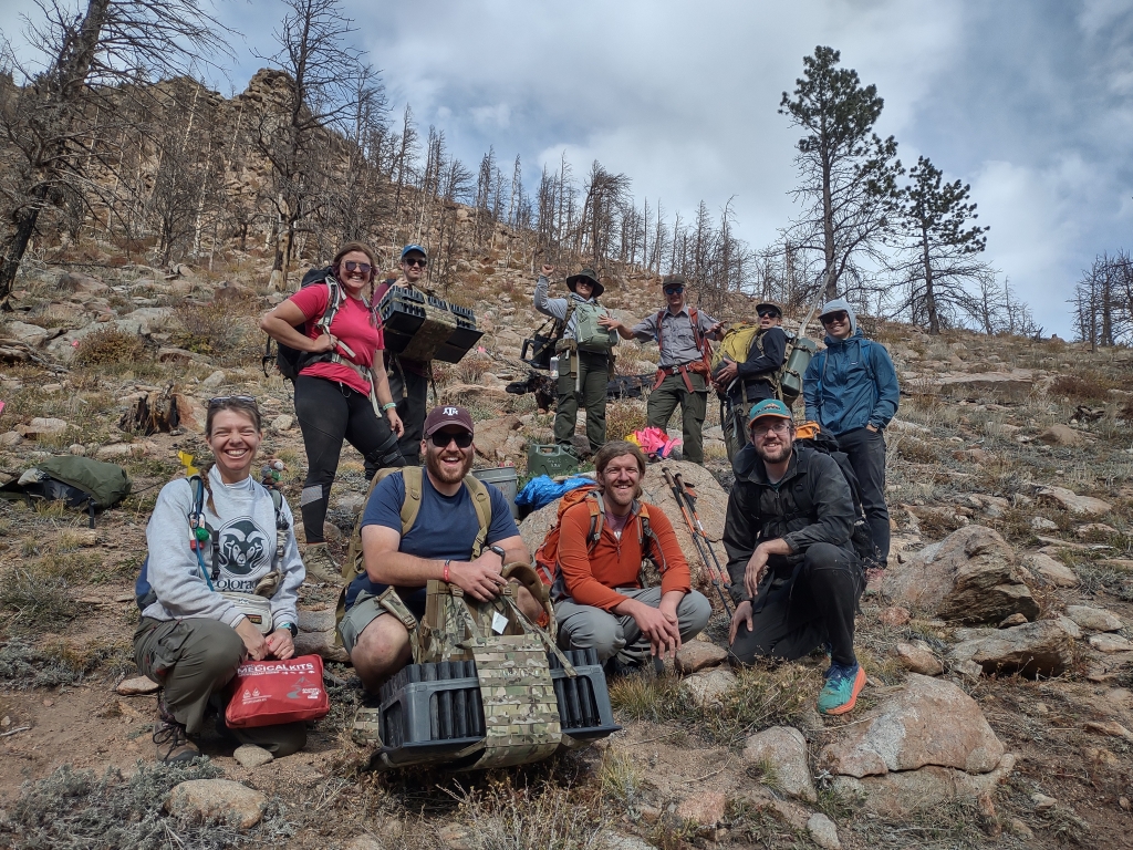 A group of hikers posing on a rocky hillside with backpacks and outdoor gear under a partly cloudy sky.