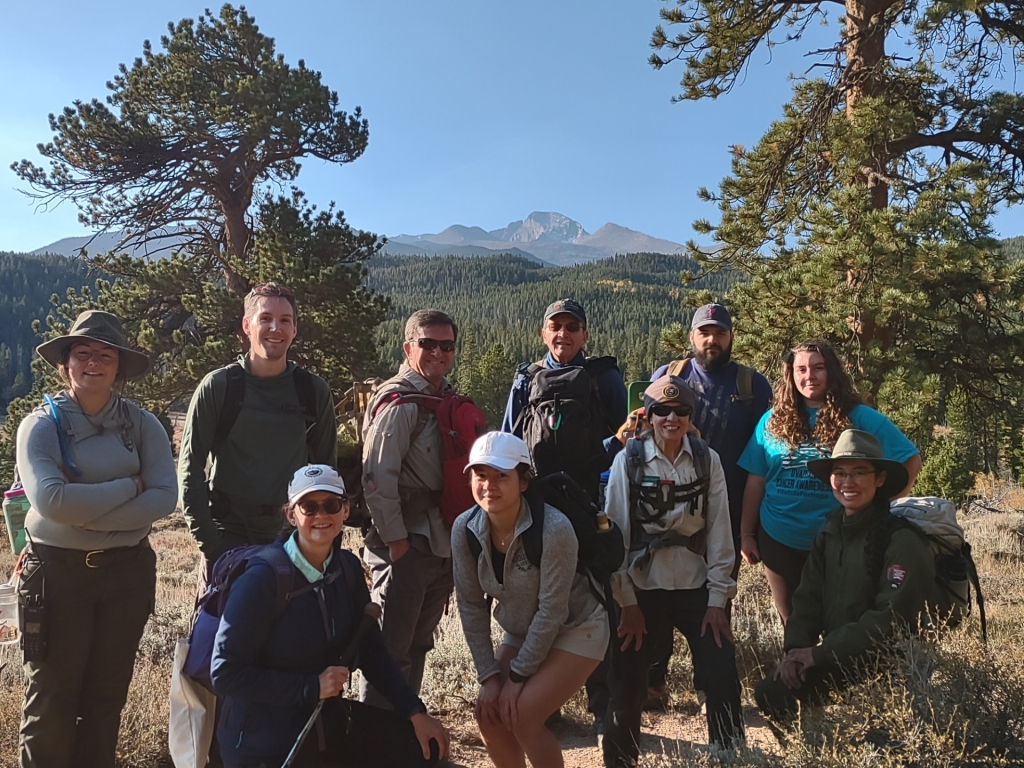 Group of hikers posing outdoors with backpacks; trees and mountain in the background under a clear blue sky.